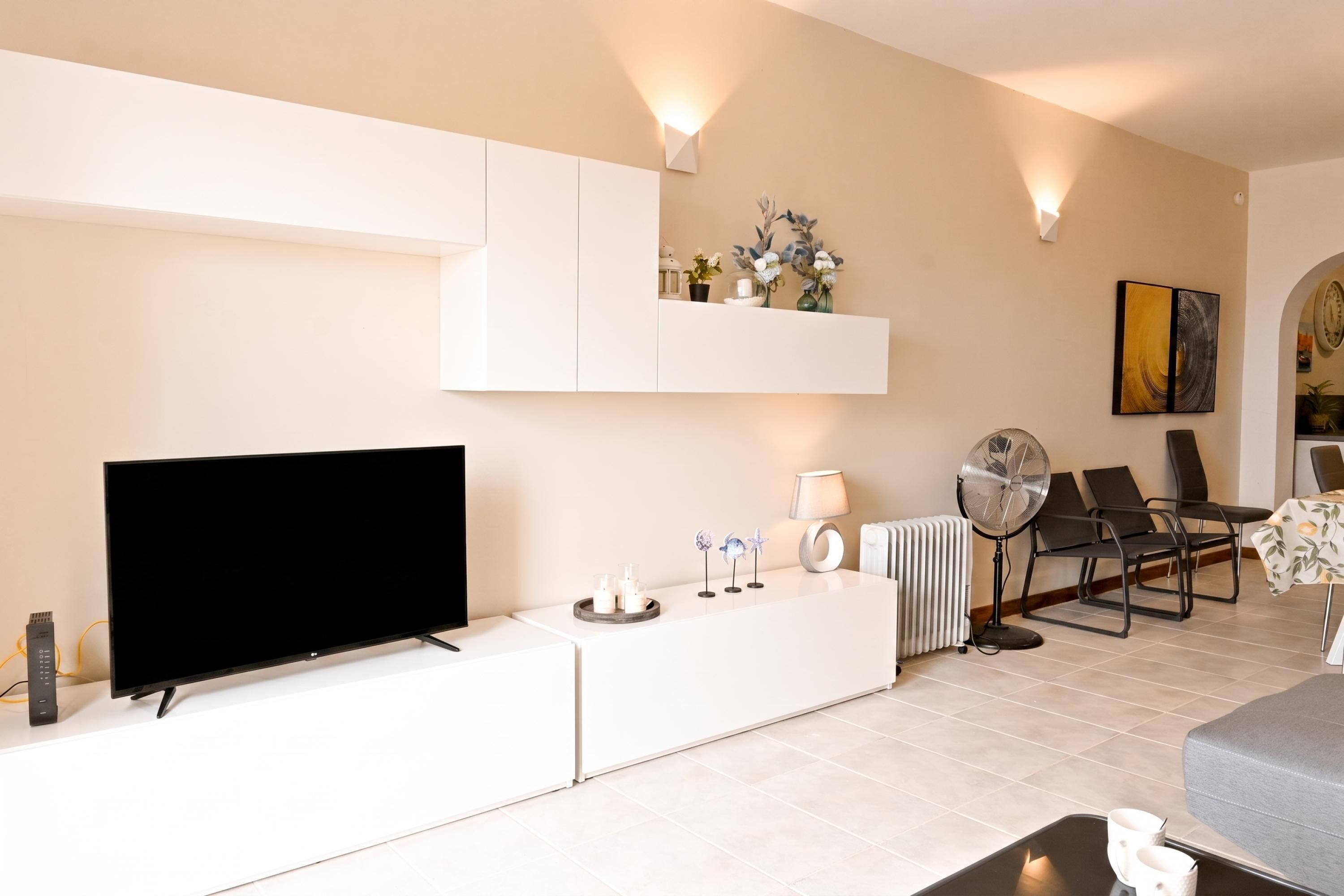 A modern living room with a flat-screen TV, a white entertainment unit, a dining area with chairs, and a radiator.