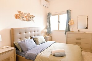 Un dormitorio con cabecera capitonada, una mesita de noche con lámpara, un aparador con cajones, una ventana con cortinas y un espejo colgado en la pared.