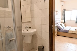 A modern bathroom with a sink, mirror, and toilet. The room features a bed with a nightstand and a lamp.