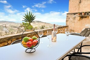 A table with a fruit bowl, wine glasses, and a menu on a terrace overlooking a landscape.