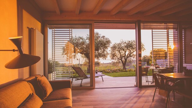An open-plan living area with a wooden floor, a sofa, a dining table with chairs, and a view of a garden through large glass doors. - San Colombano Suite (Bardolino)