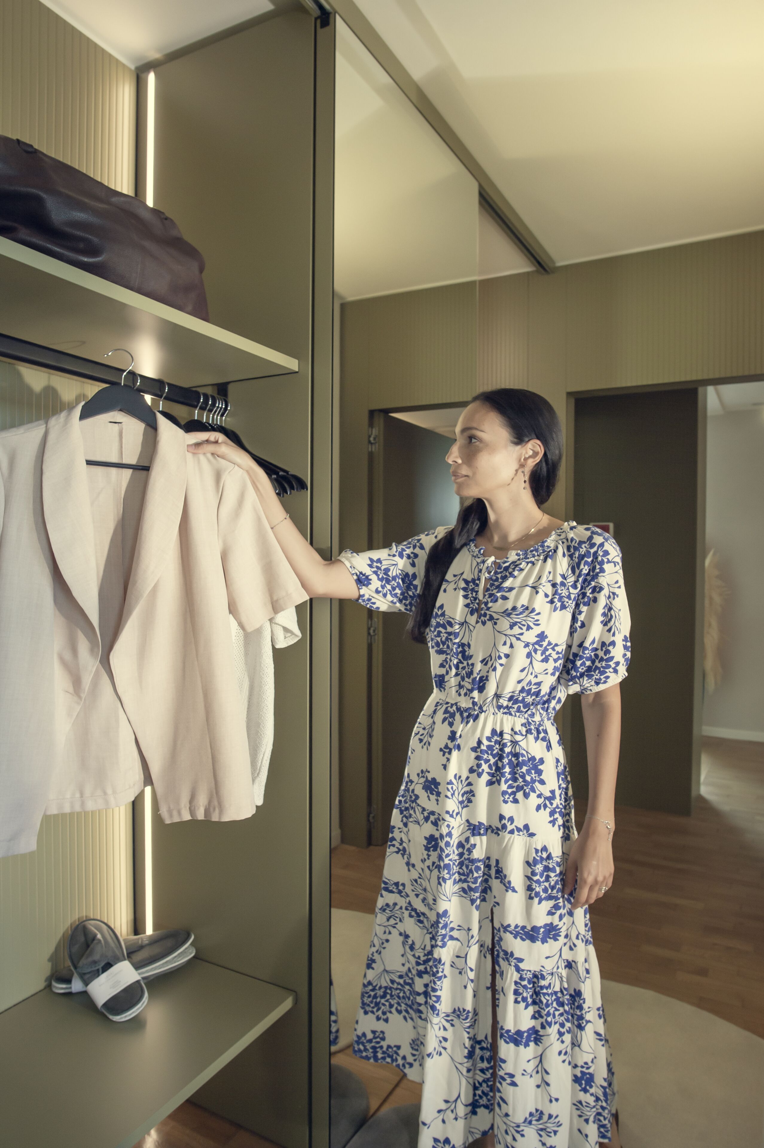 Une femme vêtue d’une robe à motifs floraux se tient dans une pièce moderne, dotée d’une armoire à portes miroir et d’un plancher en bois.