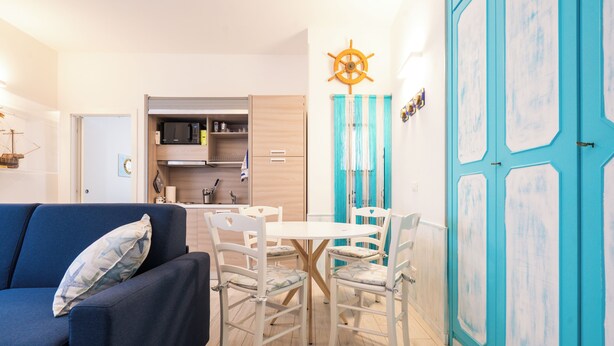 A compact living space with a blue sofa, a dining table with chairs, a kitchen area, and a large blue door.