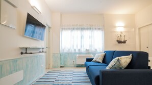 A living room with a blue sofa, a flat-screen TV mounted on the wall, a window with sheer curtains, and a decorative ship model on the wall.