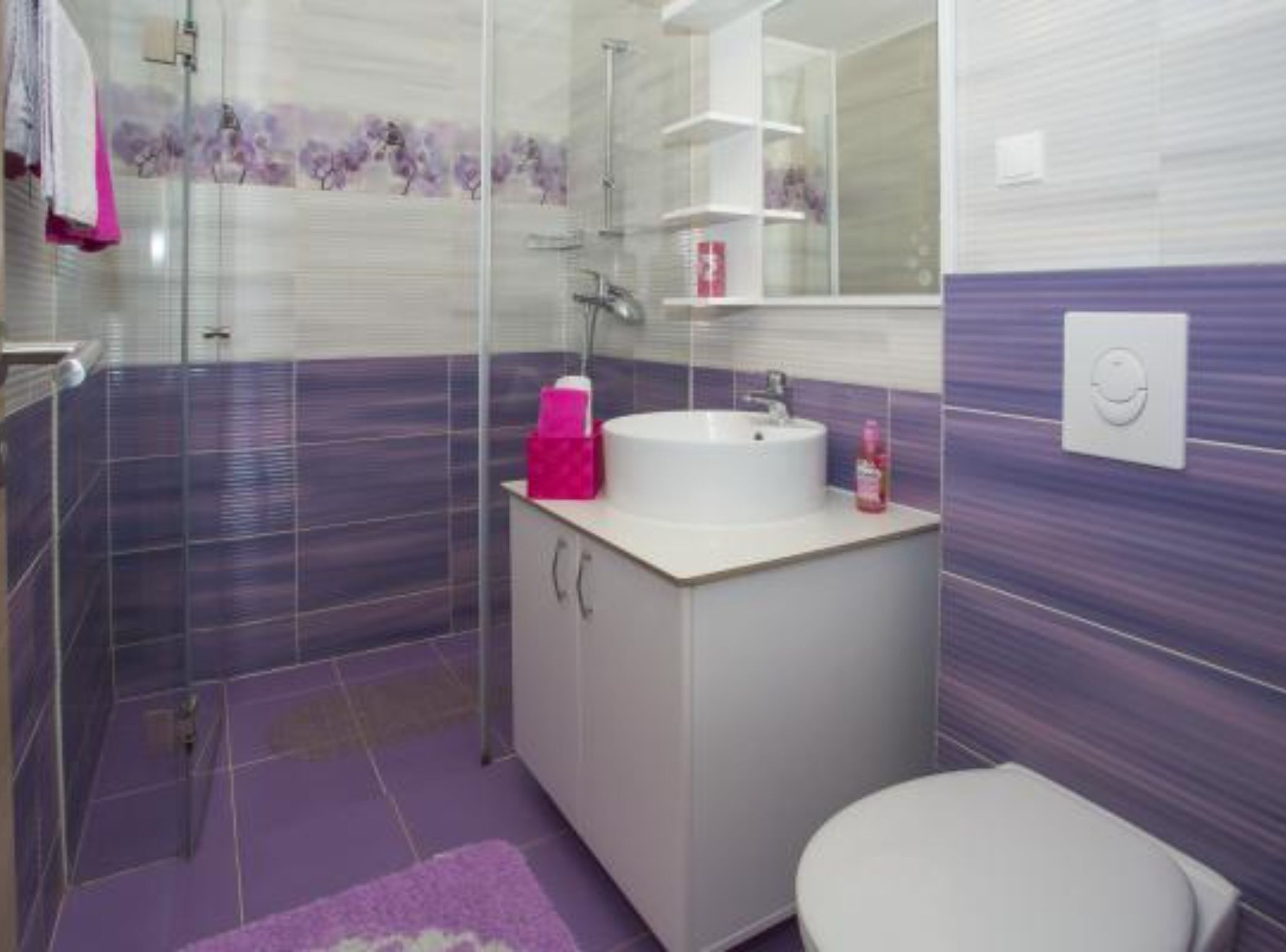 A modern bathroom with a glass shower enclosure, a white sink with a pink soap dispenser, and a purple tiled floor.