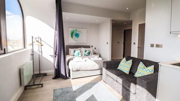 A modern bedroom with a bed, a grey sofa, a radiator, and a window with curtains. - Stunning Studio - Sleeps 4 (Staines)