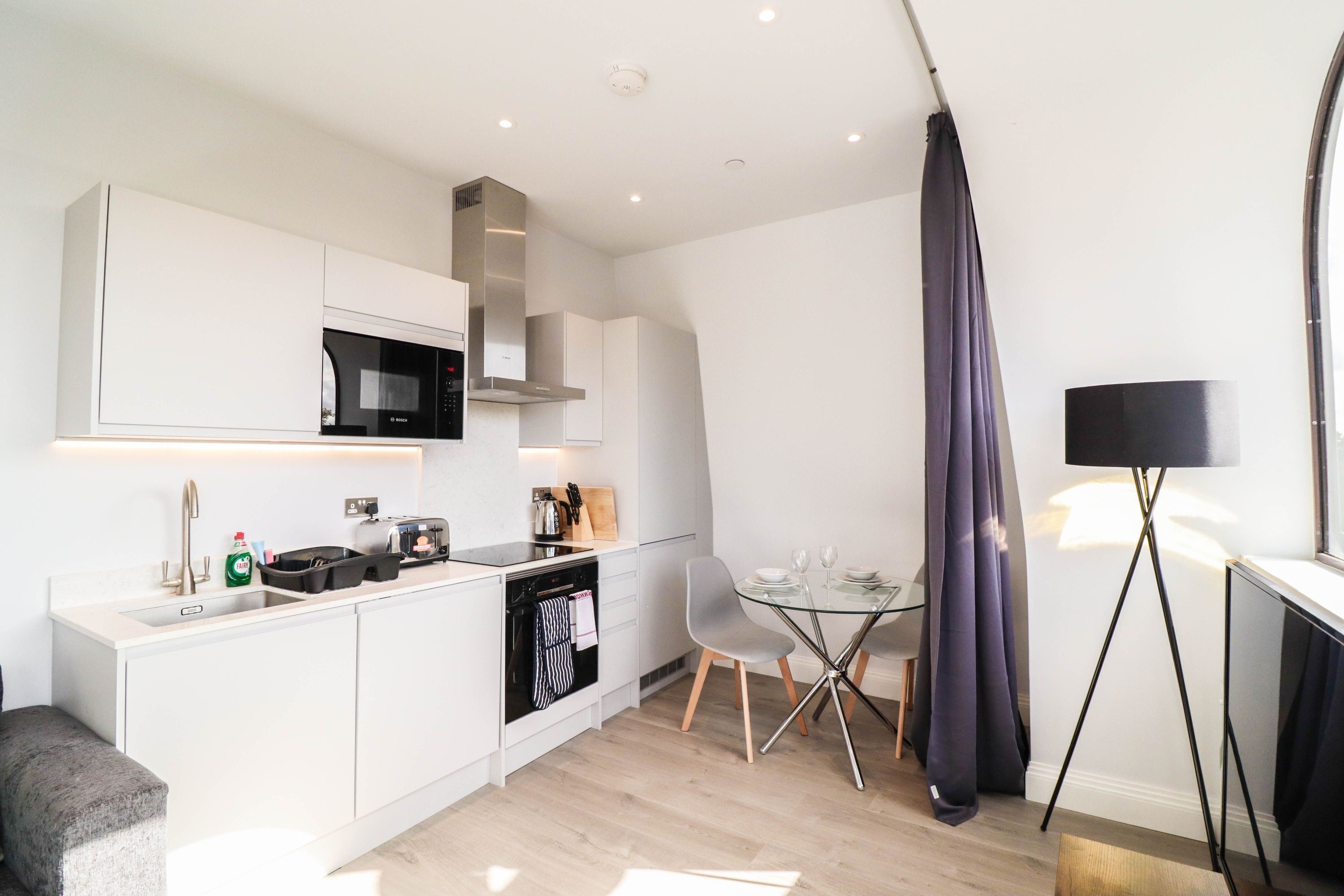 Una cocina moderna con armarios blancos, un microondas y un comedor con mesa y sillas de vidrio.