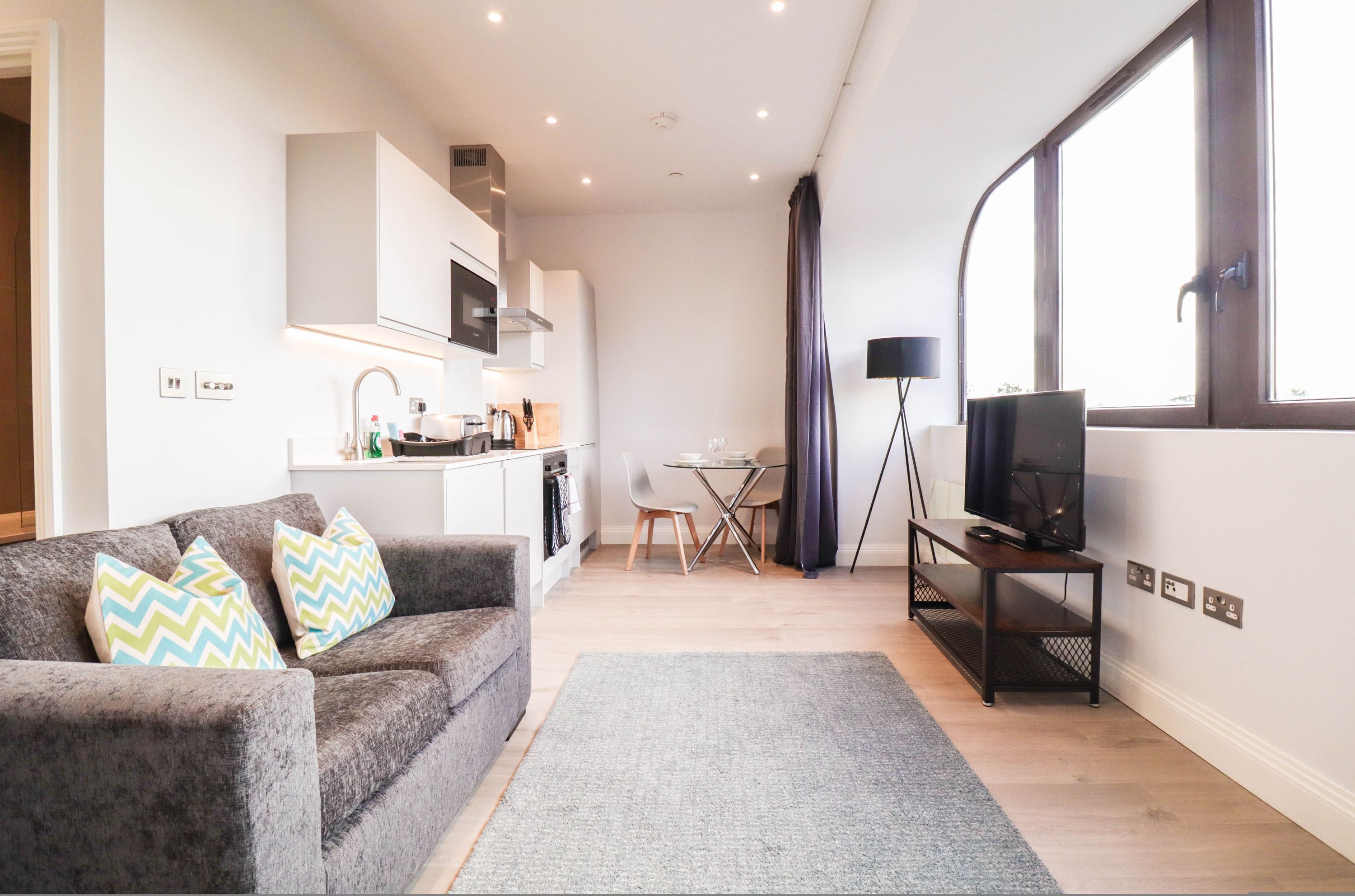 A modern living room with a grey sofa, a flat-screen TV on a stand, and a dining area with a table and chairs.