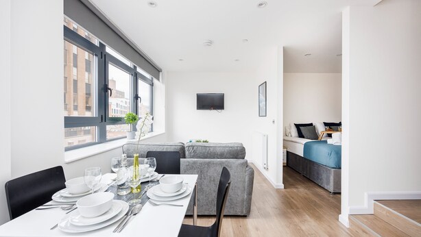 Una habitación moderna y bien iluminada con zona de comedor, un sofá gris y un televisor de pantalla plana montado en la pared.