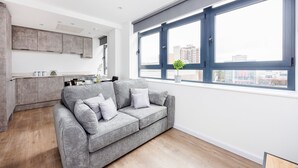 Una sala moderna con un sofá gris, una zona de cocina y ventanas amplias con vistas a la ciudad.