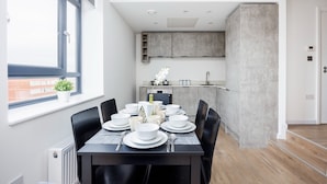 A modern dining area with a table set for six, a kitchen in the background, and a view of the city through a large window. - Modern 2 bed Flat in Town Centre - Free Wifi (Luton)