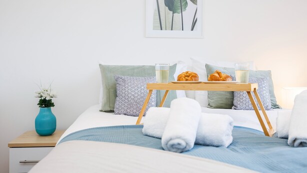 A neatly made bed with a blue and white duvet, a wooden bedside table with a vase of flowers, and a tray with croissants and a glass of milk. - Modern 2 bed Flat in Town Centre - Free Wifi (Luton)