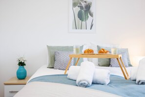 Ein ordentlich bezogenes Bett mit einer blauen und weißen Bettdecke, ein Nachttisch aus Holz mit einer Blumenvase und ein Tablett mit Croissants und einem Glas Milch.