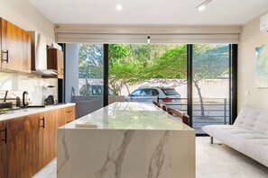 Een moderne keuken met een marmeren eiland, houten kasten en een groot raam met uitzicht op een boom en geparkeerde auto's.