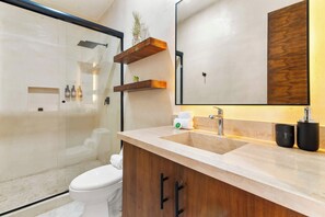 A modern bathroom with a glass shower enclosure, a toilet, a sink with a wooden cabinet, and a large mirror. - Bright Tulum 2BR - Rooftop Pool near Aldea Zama (Tulum)
