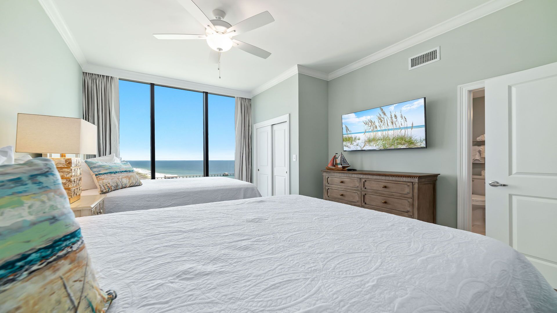 A hotel room with two beds, a ceiling fan, a large window with a view of the ocean, a flat-screen TV, and a wooden dresser.