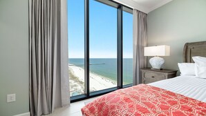 A hotel room with a large window overlooking a beach and pier, a bed with a red patterned bedspread, a nightstand with a lamp, and a wooden headboard. - New Complex!  Beachfront 4BR, Lazy River and Water Slide! (Gulf Shores)