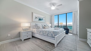 A bedroom with a large bed, a nightstand, a ceiling fan, a window with curtains, and a view of the ocean. - New Complex! Beachfront 4BR, 3RD FLOOR, Lazy River and Slide! (Gulf Shores)