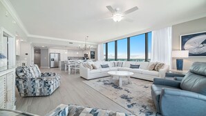 A spacious living room with a white sofa, blue armchairs, and a coffee table. There is a large window offering a view of the sky.