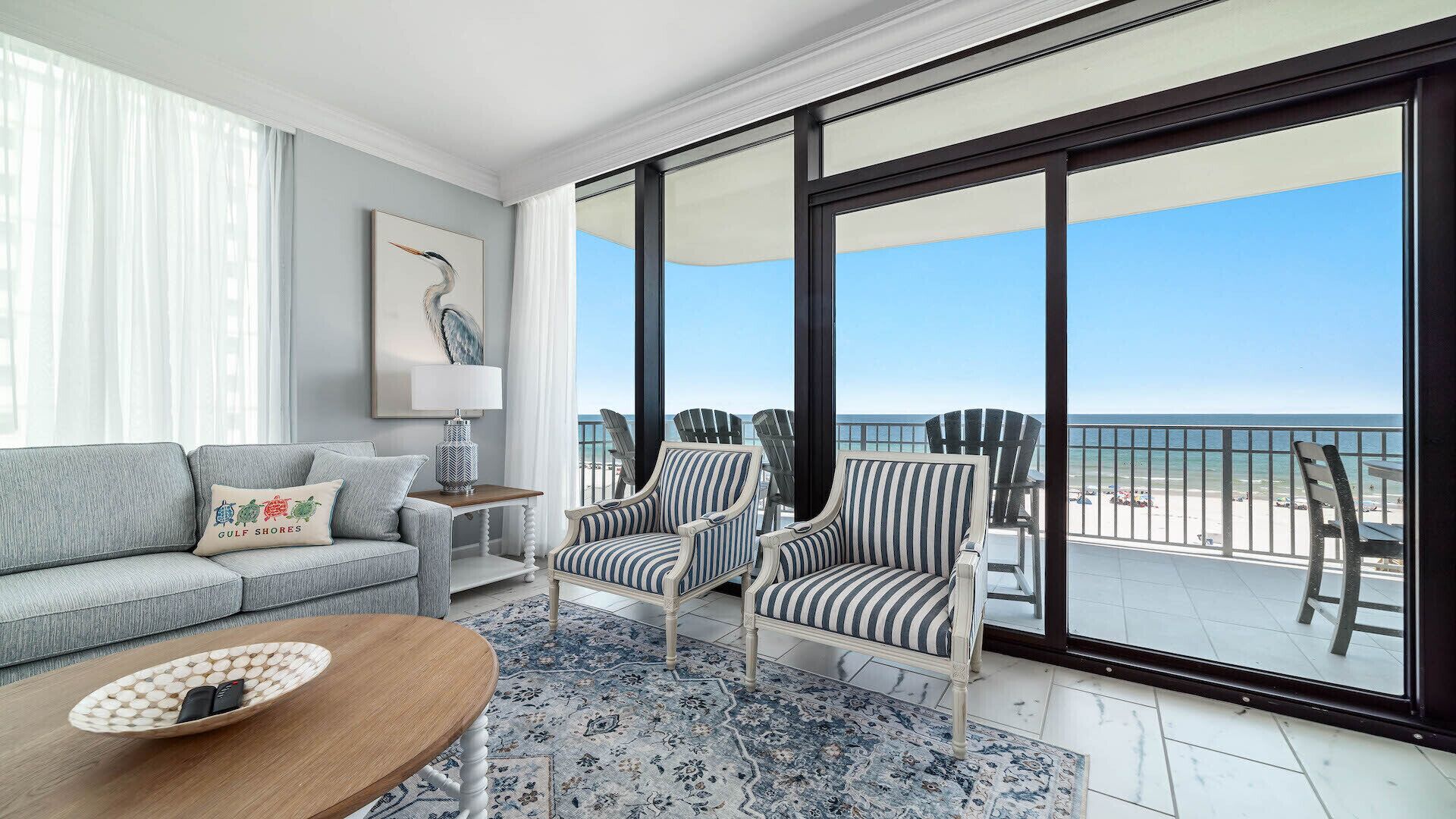 A balcony with a view of the beach, a grey sofa, striped chairs, and a table with a bowl.