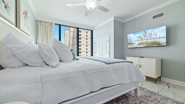 A bedroom with a bed, white bedding, a ceiling fan, a TV on the wall, and a view of a building outside.