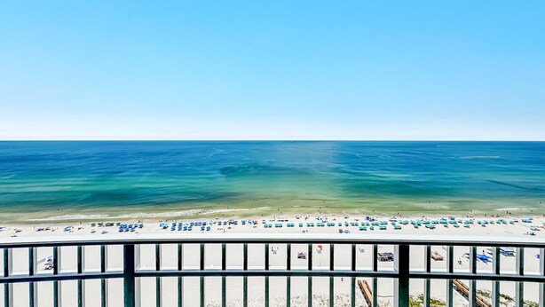 Une plage aux eaux d’un bleu limpide, une rive sablonneuse et une rambarde métallique au premier plan.