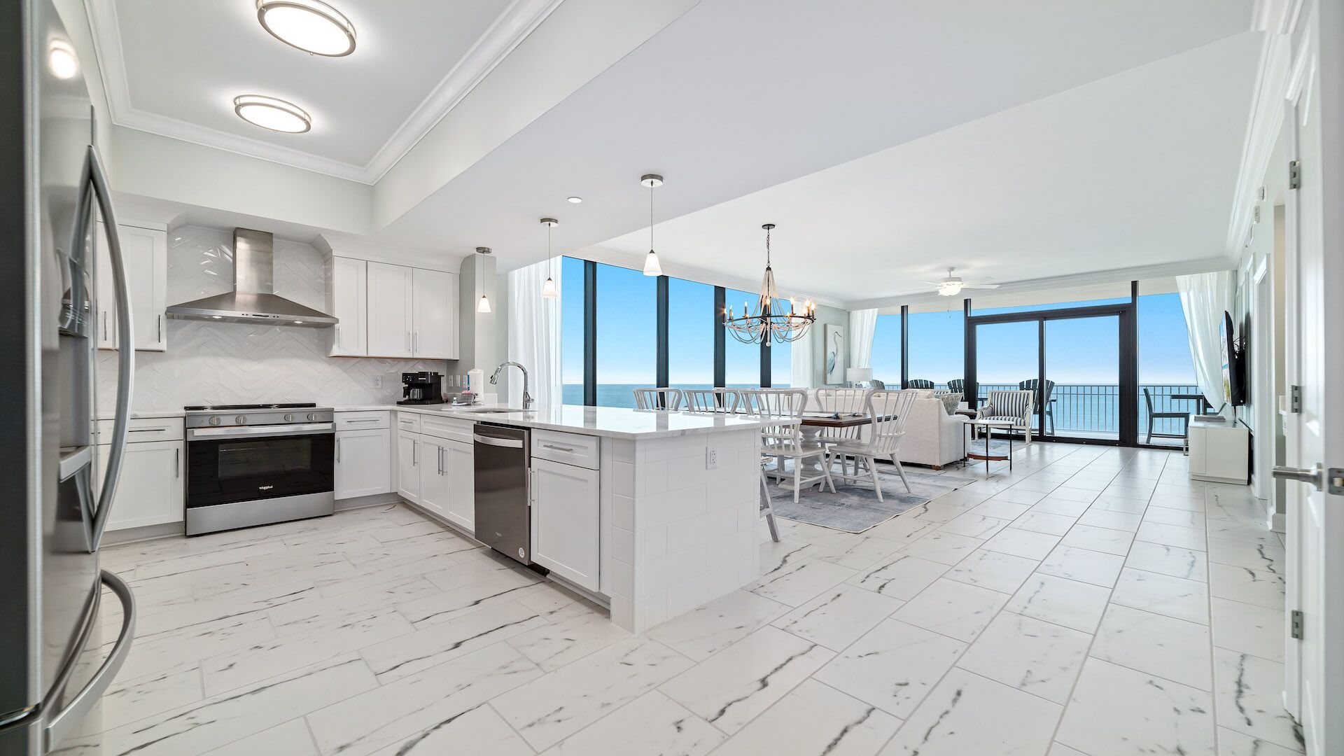 A modern kitchen with white cabinetry and a large island, a dining area with white chairs, and a balcony with a view of the ocean.