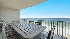 Un balcon avec une table et des chaises, donnant sur une plage où l’on aperçoit une jetée au loin.