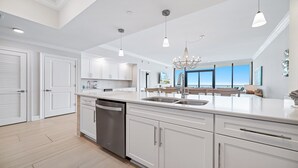 A modern kitchen with white cabinetry, a dishwasher, and a large island with a sink. There are pendant lights and a chandelier. - New Complex!  Beachfront 3BR Beauty! LAZY RIVER and WATER SLIDE!! (Gulf Shores)