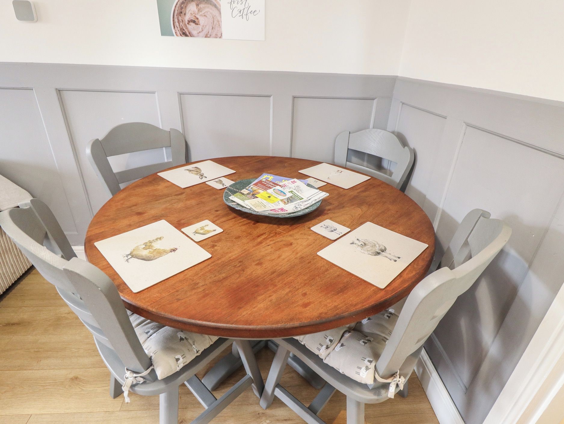 Una mesa de madera redonda con seis sillas, cada una con un cojín. Sobre la mesa, hay varias cartas y una revista.