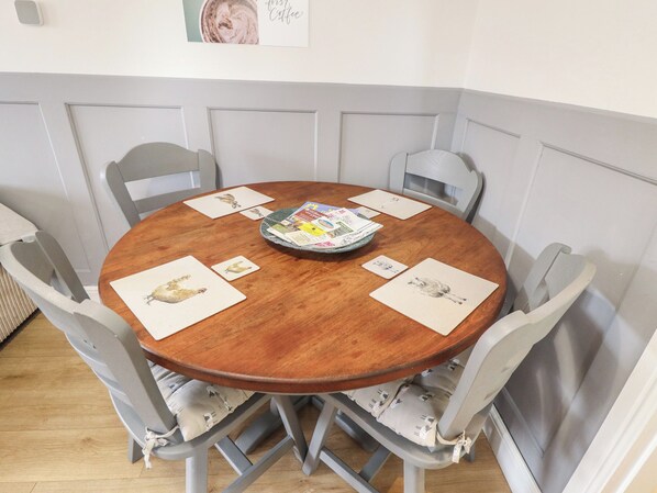 Une table de salle à manger ronde en bois, entourée de six chaises, chacune dotée d’un coussin. Sur la table, on trouve plusieurs cartes et un magazine.