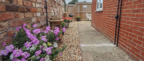 A narrow pathway leading to a brick building with a white door, surrounded by potted plants and a gravel path.