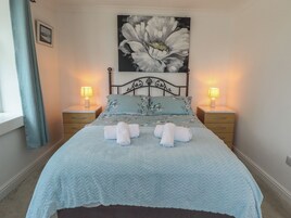 Un dormitorio con una cama, dos mesitas de noche, un buró, un cuadro floral grande y una ventana con cortinas.
