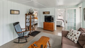 Un salon comprenant un fauteuil à bascule, une table basse en bois, un canapé marron agrémenté d’un coussin à motifs, un téléviseur posé sur un meuble en bois et une vitrine contenant des objets décoratifs.