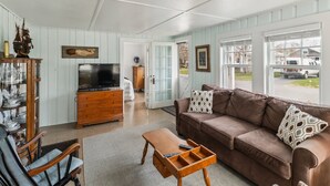 Un salon avec un canapé en cuir marron, une table basse en bois et un téléviseur posé sur un meuble en bois. Une fenêtre donne sur l’extérieur, où l’on aperçoit un camion garé.