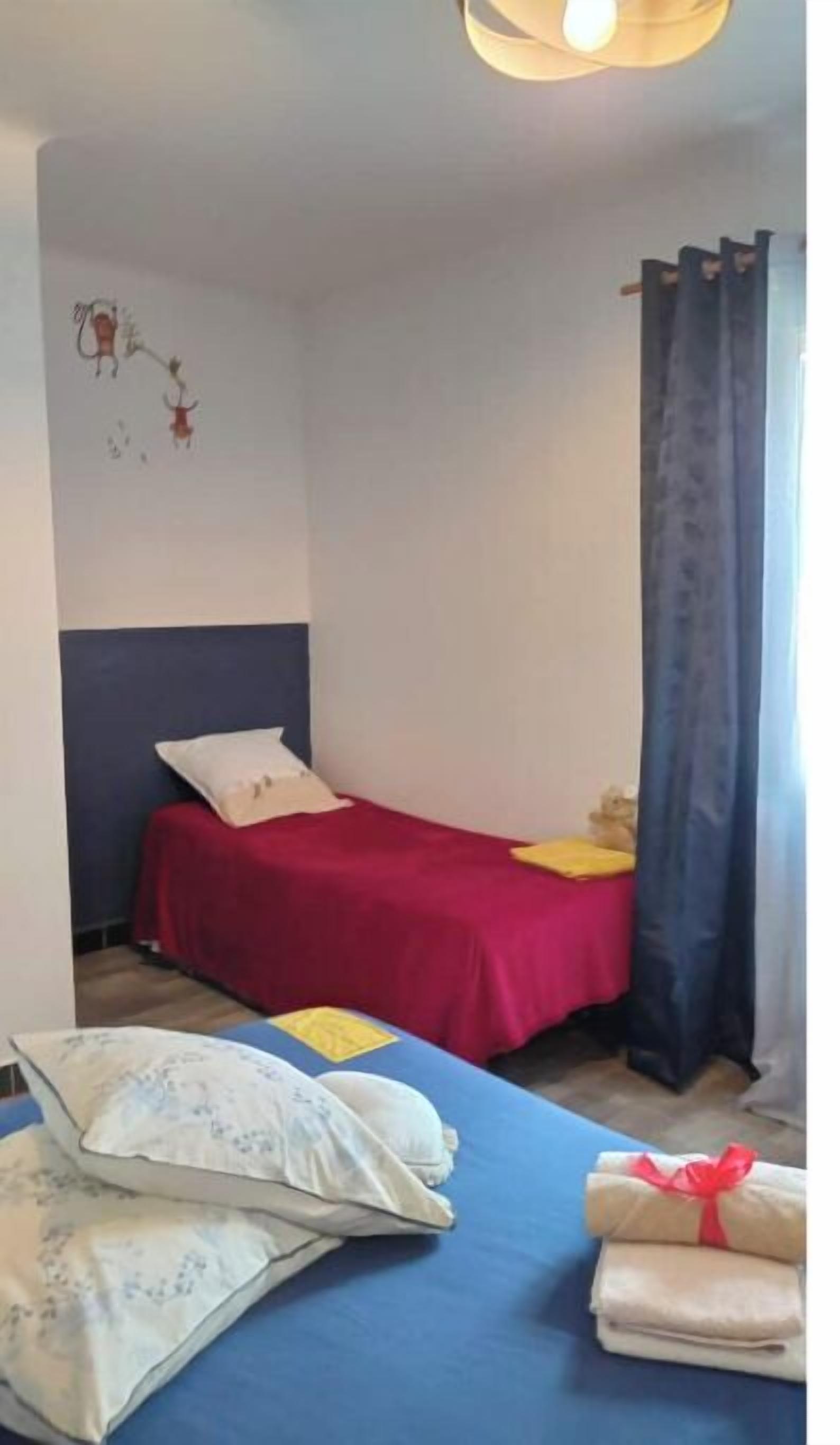 A bedroom with a single bed, a red bedspread, and a blue headboard. There are pillows and a blanket on the bed, and a curtain on the window.