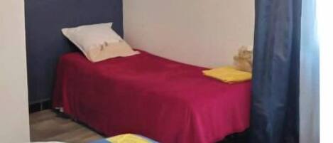 Ein Schlafzimmer mit einem Einzelbett, einer roten Bettdecke und einem blauen Kopfteil. Auf dem Bett liegen Kissen und eine Decke, und am Fenster hängt ein Vorhang.