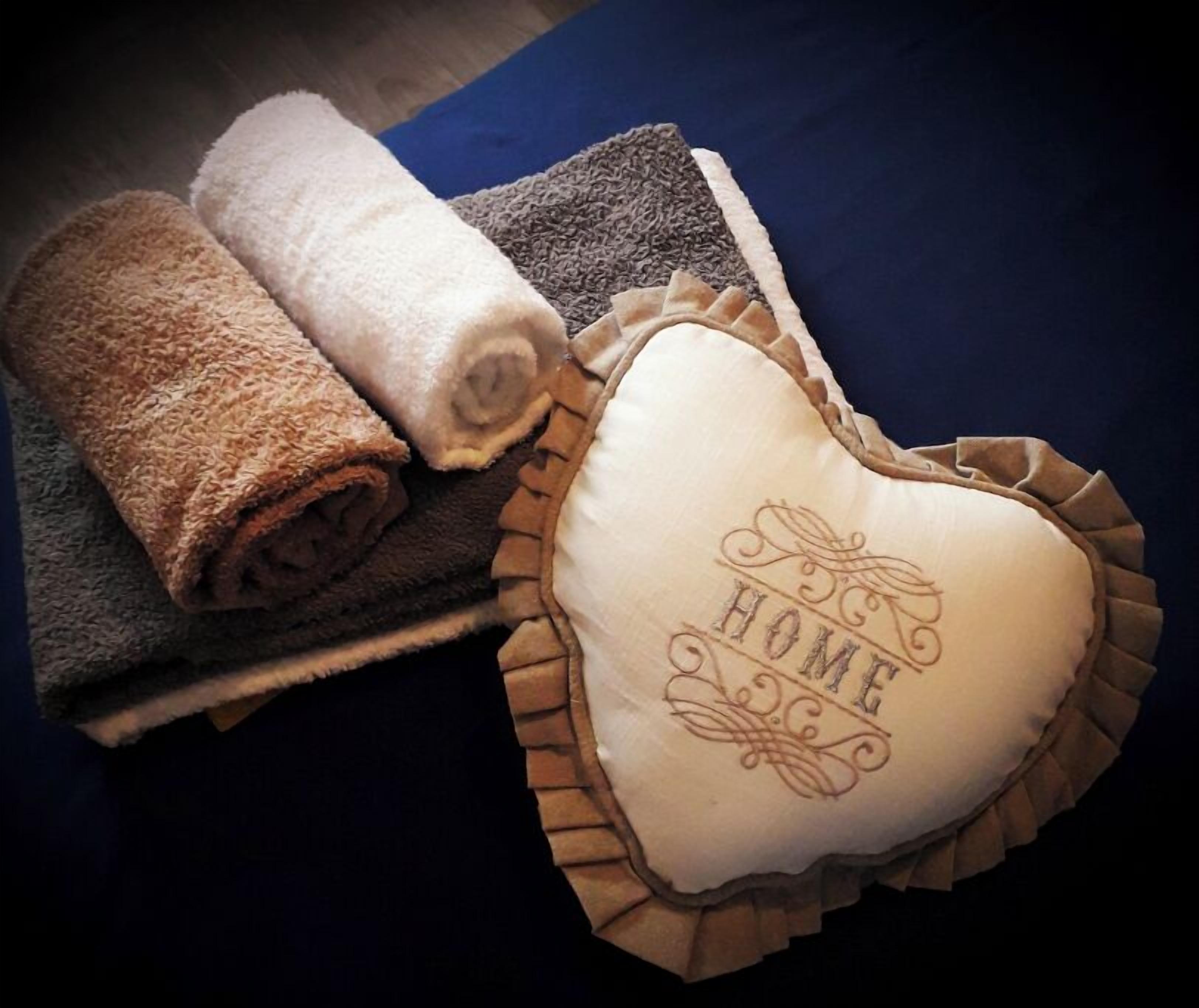 A rolled towel and a heart-shaped pillow with "HOME" embossed on it, placed on a dark blue bedspread.