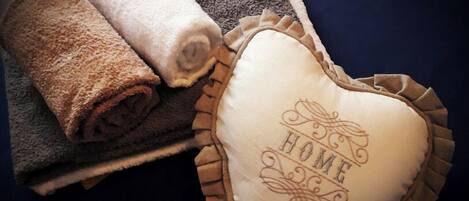 A rolled towel and a heart-shaped pillow with "HOME" embossed on it, placed on a dark blue bedspread.