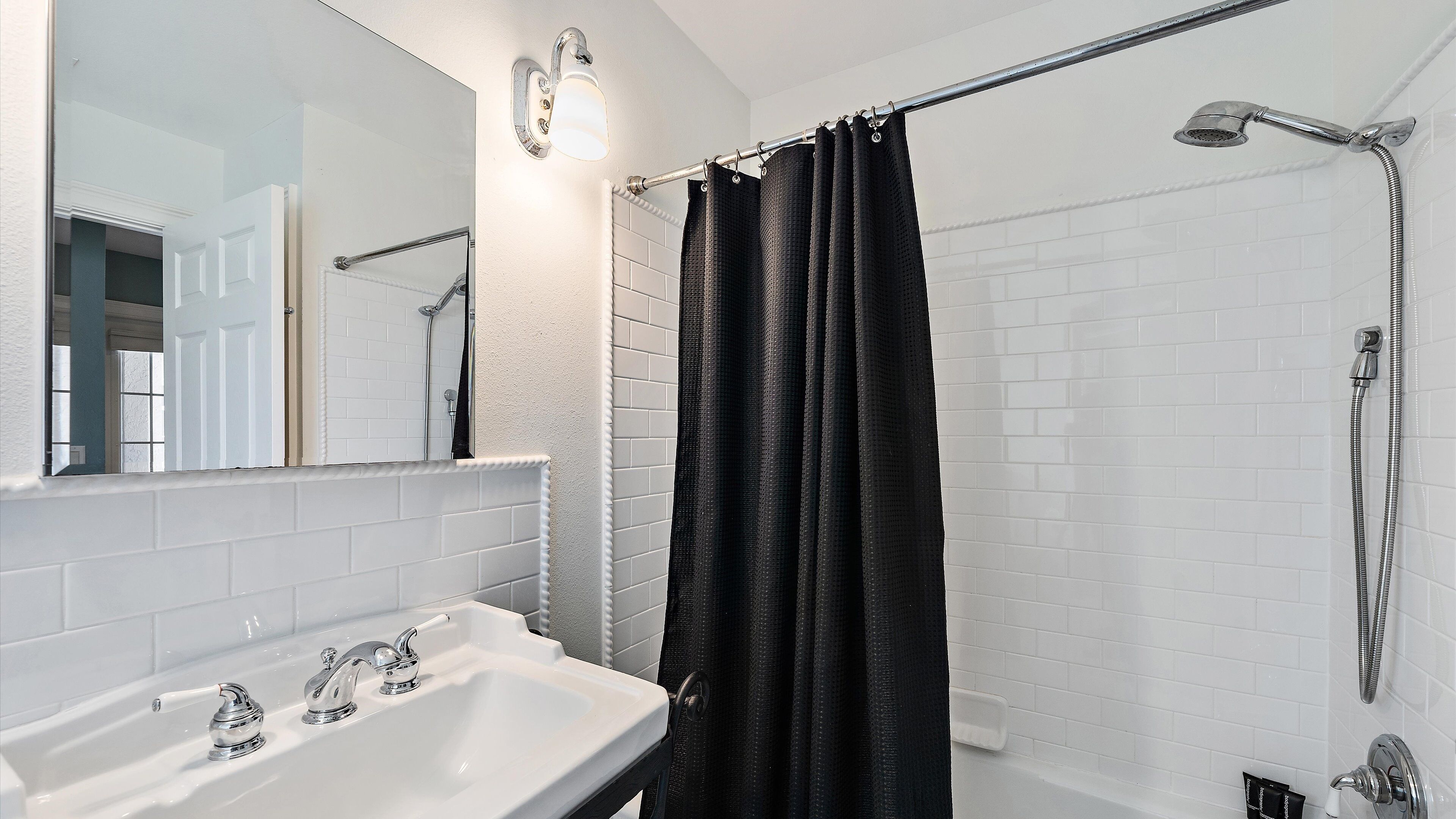 Une salle de bain avec un lavabo blanc, un miroir, une douche avec un pommeau de douche amovible et un rideau de douche noir.