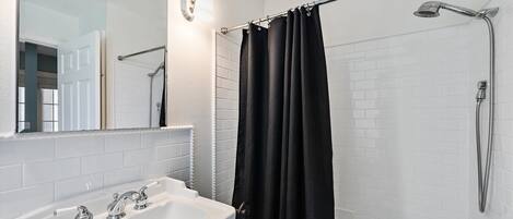Une salle de bain avec un lavabo blanc, un miroir, une douche avec un pommeau de douche amovible et un rideau de douche noir.