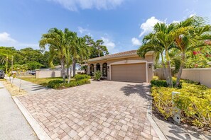 A residential property with a paved driveway, a garage, and a well-maintained garden with palm trees and shrubs. - Sapphire Shores 3 Bedroom Home (Sarasota)