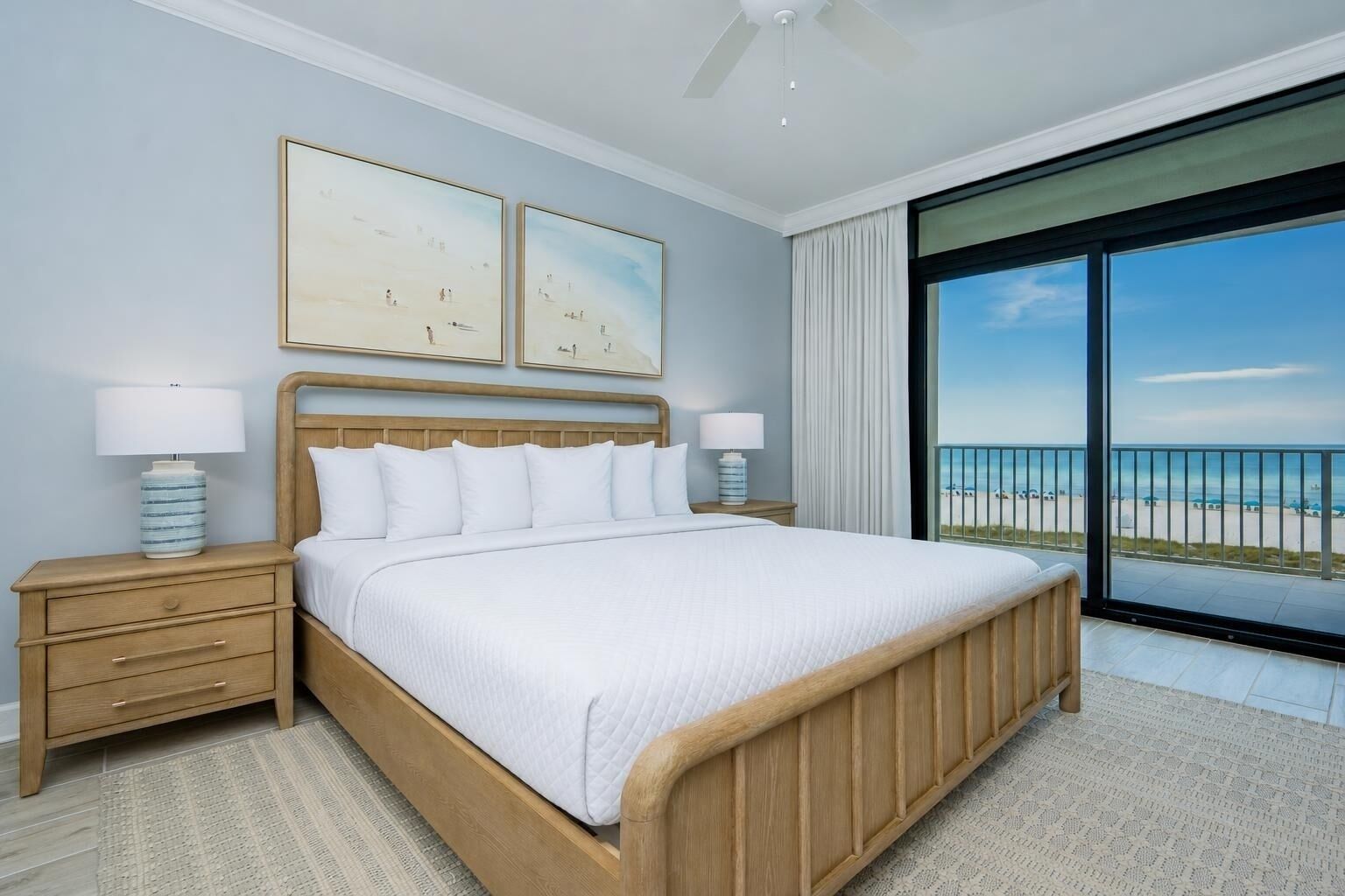 A bedroom with a large bed, two bedside tables, a ceiling fan, and a balcony with a view of the beach.