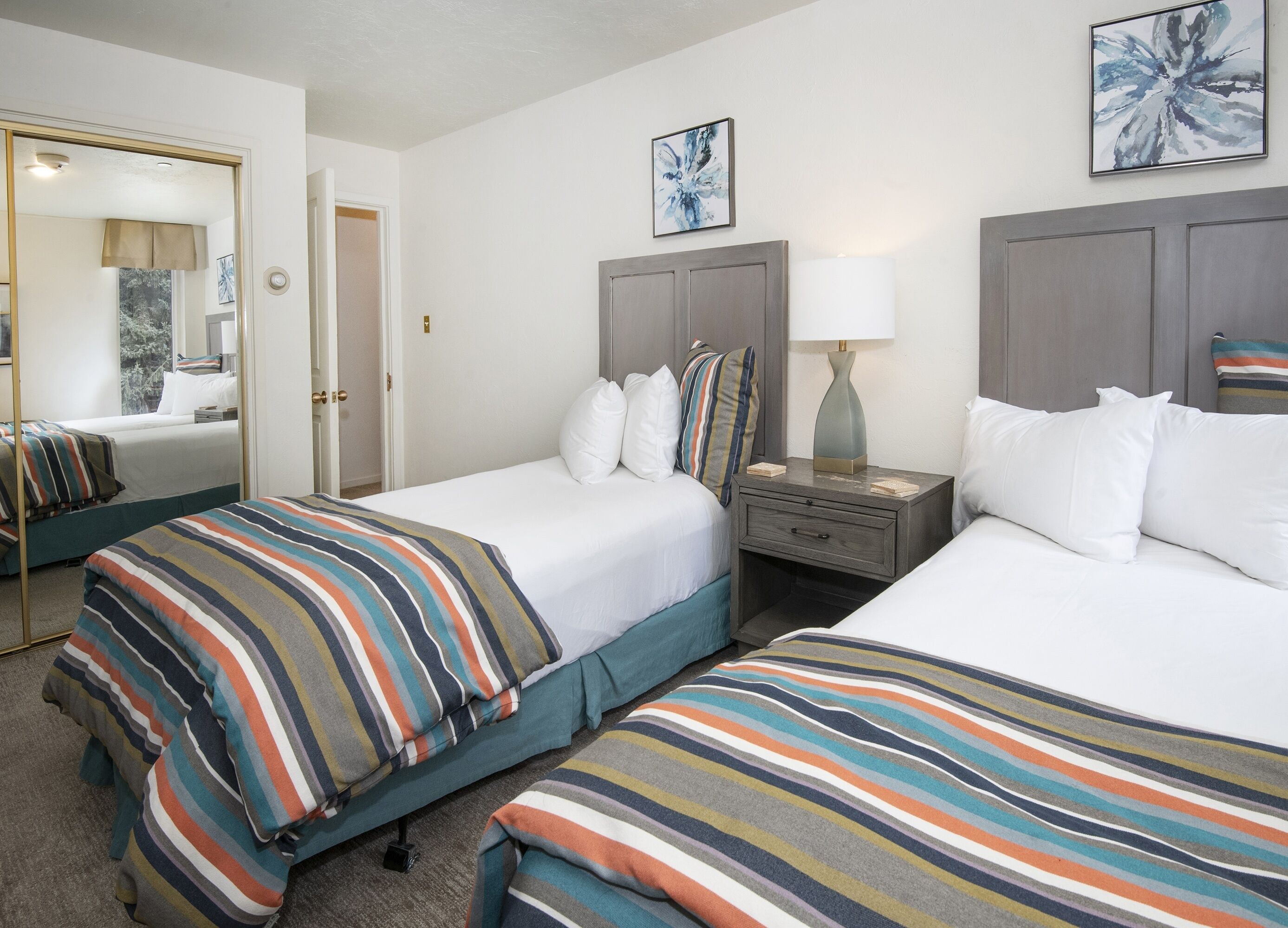 Habitación de hotel con dos camas, una mesita de noche con lámpara, un espejo y una puerta que da a otra habitación.