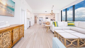 A spacious living area with a large window, a white sofa, a wooden coffee table, and a dining table with chairs. - Phoenix Gulf Shores Ii 705 4 Bedroom Condo (Gulf Shores)