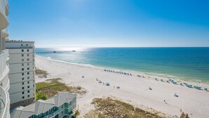 A beach view with a pier extending into the ocean, a sandy beach with umbrellas, and a high-rise building in the foreground. - Phoenix Gulf Shores Ii 1102 3 Bedroom Condo (Gulf Shores)