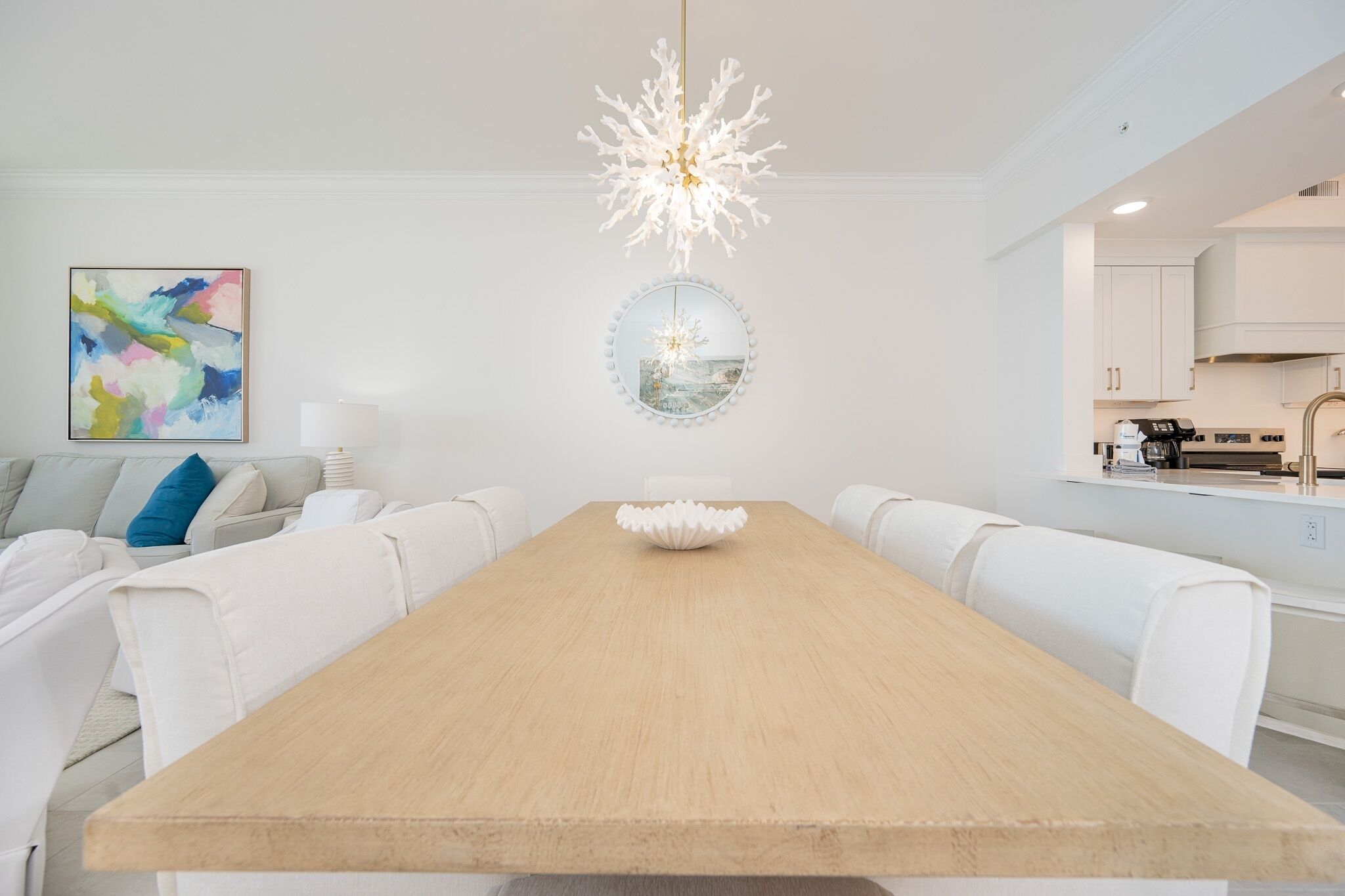 Ein zeitgemäßer Essbereich mit Holztisch, weißen Stühlen, einer Chandelierleuchte, einem Gemälde und einer Küche im Hintergrund.