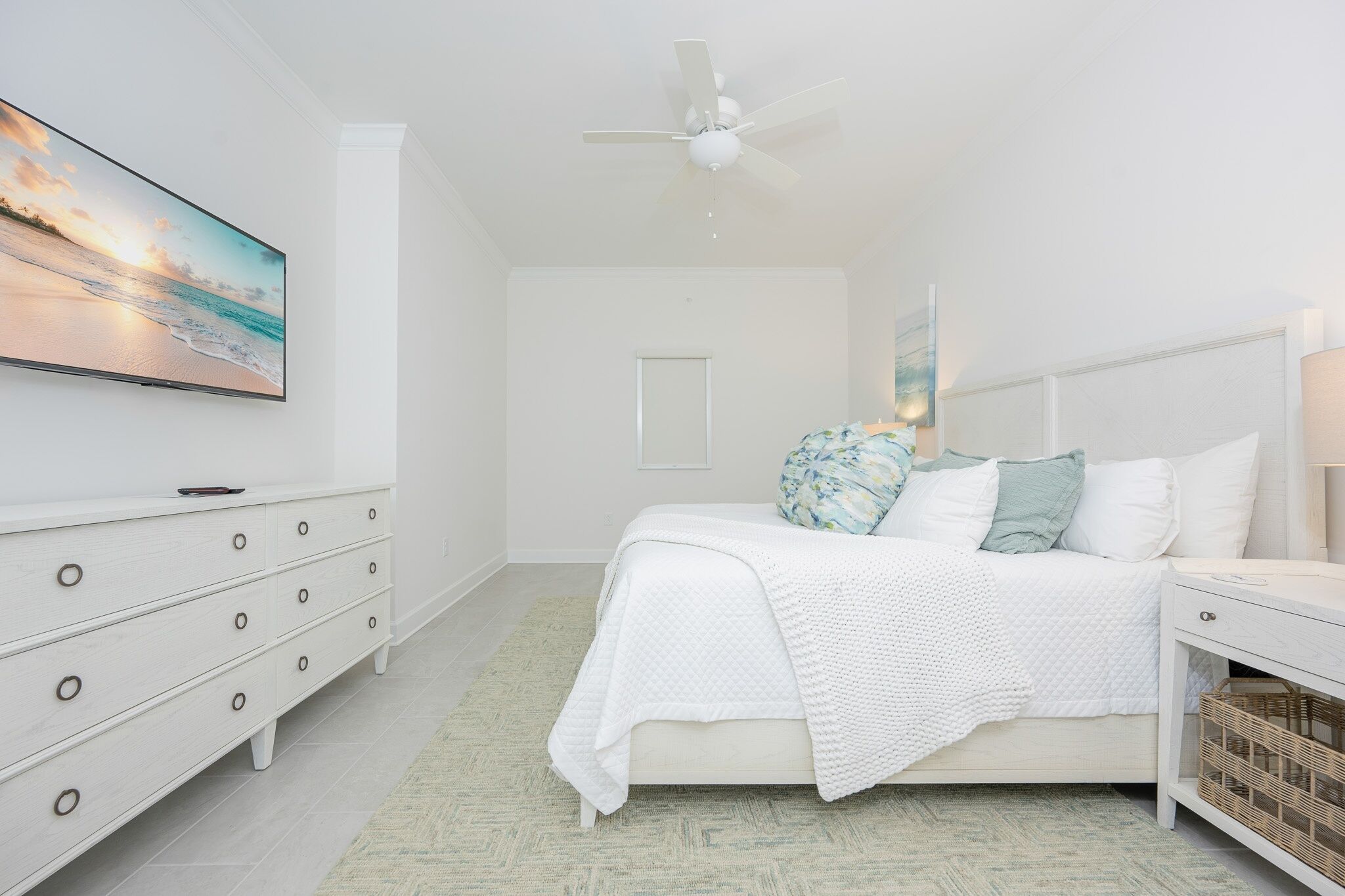 A bedroom with a bed, a dresser, a nightstand, a ceiling fan, and a wall-mounted TV displaying a beach scene.