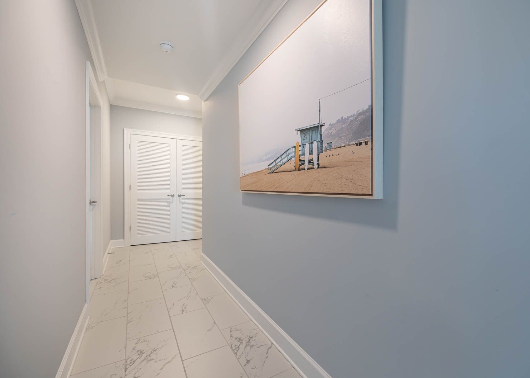 A hallway with a white door, a framed beach scene on the wall, and a tiled floor.