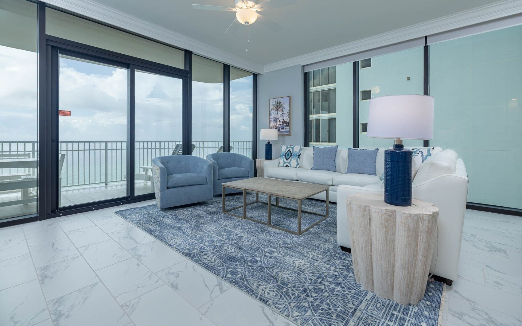 A spacious living room with a large sliding glass door leading to a balcony, a white sofa, two armchairs, a coffee table, and a lamp on a wooden side table.